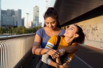 Две молодые женщины с длинными каштановыми волосами стоят на городском мосту, обнимаются и улыбаются. — стоковое фото