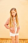 Portrait of girl with long blond hair wearing shorts, shirt and pink jacket, on pale yellow background. — Stock Photo