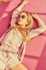 Porträt eines Mädchens mit langen blonden Haaren, auf dem Rücken liegend, Sonnenbrille, Shorts und Jacke tragend, auf rosa Hintergrund. — Stockfoto