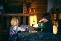 Junge und Mädchen sitzen an einem Tisch in einem Blockhaus und spielen Schach, Vasterbottens Lan, Schweden. — Stockfoto