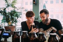 Bearded young man with brown hair and tattoos and young woman with short hair sitting in a bar, looking at mobile phone. — Stock Photo