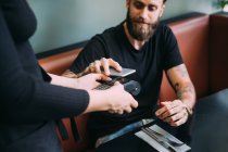 Alto ángulo de primer plano del hombre barbudo tatuado sentado en un bar, utilizando el teléfono móvil para pagar. - foto de stock