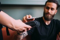 Close up of tattooed bearded man sitting in a bar, using mobile phone to pay. — Stock Photo