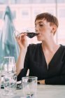 Mujer joven con el pelo corto con top negro sentado en la mesa en un bar, bebiendo vino tinto. - foto de stock