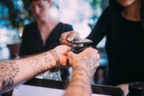 Close up of tattooed bartender standing at counter, handing card reader to woman using mobile phone to pay. — Stock Photo