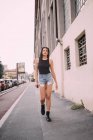 Portrait of young woman with dark brown hair, wearing black vest and denim shorts, walking down street. — Stock Photo