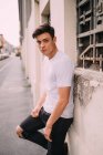 Portrait of young man with dark brown hair, wearing white T-shirt and ripped black jeans, leaning against wall. — Stock Photo