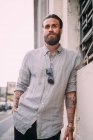 Portrait of bearded young man with brown hair, with tattoos on arms, wearing grey shirt, leaning against wall. — Stock Photo