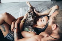 Homem tatuado barbudo com longos cabelos morena e mulher com longos cabelos castanhos abraçando com gato cinza fofo em um sofá. — Fotografia de Stock