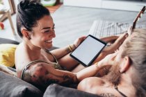 Бородатий татуйований чоловік з довгим брюнетка волоссям і жінка з довгим коричневим волоссям сидить на дивані, дивлячись на цифровий планшет . — стокове фото