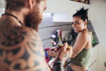 Hombre tatuado barbudo con el pelo largo morena y mujer con el pelo largo marrón de pie en una cocina, sonriendo el uno al otro. - foto de stock