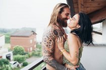Bearded tattooed man with long brunette hair and woman with long brown hair standing on a balcony, hugging and kissing. — Stock Photo