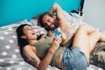 Bearded tattooed man with long brunette hair and woman with long brown hair lying on a bed, laughing. — Stock Photo