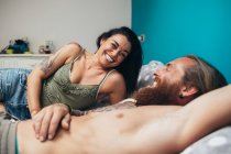Bearded tattooed man with long brunette hair and woman with long brown hair lying on a bed, smiling at each other. — Stock Photo