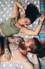 Homme tatoué barbu aux longs cheveux bruns et femme aux longs cheveux bruns allongés sur un lit, se souriant. — Photo de stock