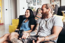 Бородатий татуйований чоловік з довгим волоссям брюнетки і жінка з довгим коричневим волоссям сидить на дивані, посміхаючись, граючи в консольну гру . — стокове фото
