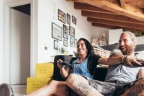 Homem tatuado barbudo com cabelos longos morena e mulher com cabelos longos marrom sentado em um sofá, sorrindo enquanto joga o jogo de consola. — Fotografia de Stock