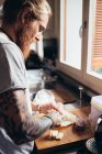 Barbudo hombre tatuado con el pelo largo morena de pie en una cocina, la preparación de alimentos. - foto de stock