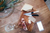 High angle close up of knives, smoked ham and hard cheese on wooden cutting board. — Stock Photo