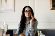 Woman with long black hair wearing glasses sitting at a restaurant table, using mobile phone. — Stock Photo