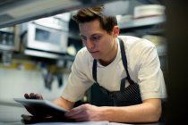 Chef vistiendo delantal azul de pie en la cocina, utilizando tableta digital. - foto de stock