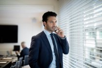 Бородатий бізнесмен в костюмі і краватці стоїть біля вікна, використовуючи мобільний телефон . — стокове фото