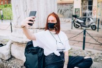 Junge Frau trägt Gesichtsmaske während Coronavirus, sitzt im Freien und macht Selfie. — Stockfoto