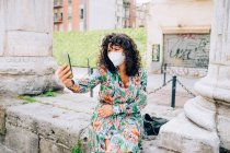Jeune femme portant un masque facial pendant le virus Corona, assise à l'extérieur, prenant selfie. — Photo de stock