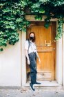 Junge Frau mit Gesichtsmaske während Coronavirus, steht vor Haustür und blickt in Kamera. — Stockfoto