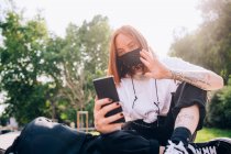 Молода жінка в масці для обличчя під час вірусу Корони, сидить на відкритому повітрі, перевіряє мобільний телефон . — стокове фото