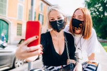 Дві молоді жінки, одягнені в маски для обличчя під час вірусу Корони, сидять на березі річки, приймаючи селфі . — стокове фото