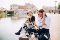 Троє молодих жінок сидять на березі річки, п'ють і використовують мобільний телефон . — стокове фото