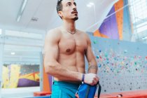 Bare-chested muscular man standing in front of indoor rock climbing wall. — стоковое фото