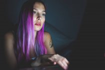Giovane donna con lunghi capelli rosa guardando un computer portatile, viso illuminato dallo schermo bagliore. — Foto stock