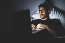 Barbudo joven acostado en la cama por la noche, mirando a la pantalla del ordenador portátil - foto de stock