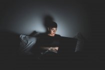 Mujer joven con el pelo corto acostado en la cama por la noche, mirando a la computadora portátil. - foto de stock