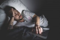 Bearded young man lying in bed at night, looking at laptop screen monitor — Stock Photo