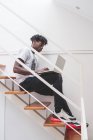 Young man with short dreadlocks sitting on staircase, typing on laptop notebook — Stock Photo