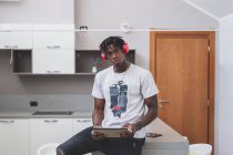 Young man with short dreadlocks sitting in kitchen, wearing headphones and holding digital tablet, looking at camera — Stock Photo