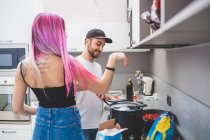 Young woman with long pink hair and bearded man wearing baseball cap standing in a kitchen, cooking food meal — Stock Photo