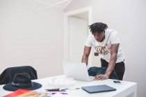Young African american man with short dreadlocks standing at table, looking at laptop notebook — Stock Photo