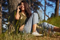 Portrait of young woman with long brown hair sitting under a tree in a forest, talking on mobile phone — Stock Photo