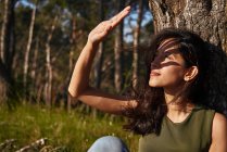 Portrait of young woman sitting under a tree in a forest, shading her eyes from the sun — Stock Photo