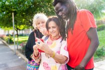 Schwarzer Mann mit Dreadlocks und zwei kaukasische Frauen stehen auf Brücke und schauen auf Handy. — Stockfoto