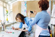 Artist painting on fabric in creative studio, mature woman photographing her using smartphone — Stock Photo