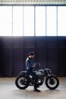 Young male motorcyclist straddling vintage motorcycle in empty warehouse, full length — Stock Photo