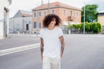 Smiling man with tattooed arms and long brown curly hair walking down street. — Stock Photo