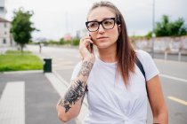 Ritratto di donna con lunghi capelli castani e braccio tatuato, indossando maglietta bianca e occhiali, utilizzando il telefono cellulare. — Foto stock