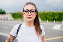 Portrait of woman with long brown hair and tattooed arm, wearing white T-Shirt and glasses, looking at camera. — Stock Photo