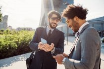 Two businessmen wearing suits standing outdoors, checking their mobile phones. — Stock Photo